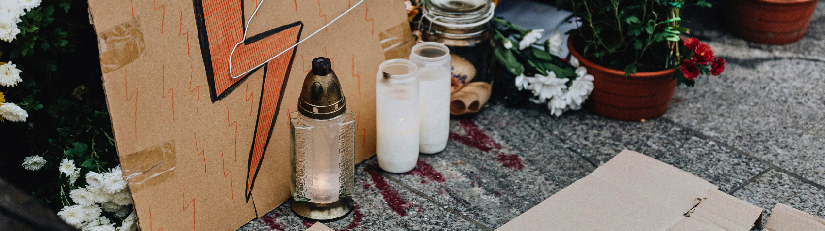 This image depicts a makeshift memorial or tribute display set up on a concrete surface, likely a sidewalk. The scene includes a cardboard sign with a lightning bolt design, a glass candle or lantern, two white cylindrical containers (possibly candles or vases), and potted flowers with white and red blooms. White flower petals are scattered on the ground, and there's a small piece of paper or card visible. Notably, there appear to be red stains or marks on the concrete. Protest-style imagery (the lightning bolt sign) with memorial elements (flowers, candles).