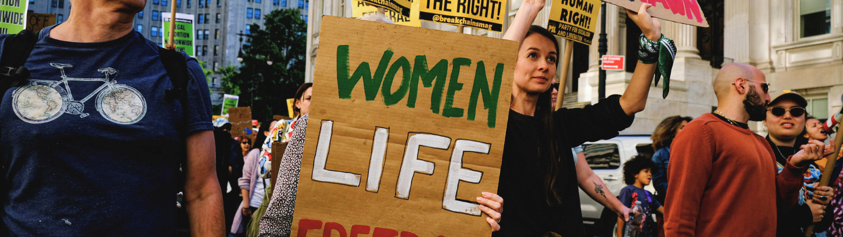 This image shows a protest or demonstration taking place on a city street. In the foreground, a woman with long dark hair is holding up a large cardboard sign that reads "WOMEN LIFE" in green and white letters. Around her are other protesters holding various signs, including ones mentioning "human rights" and "the right." To the left, there's a person wearing a blue t-shirt with a bicycle design. The background shows urban buildings and trees, indicating this is likely occurring in a downtown area. The scene appears crowded with many people participating in what seems to be a women's rights or social justice rally.