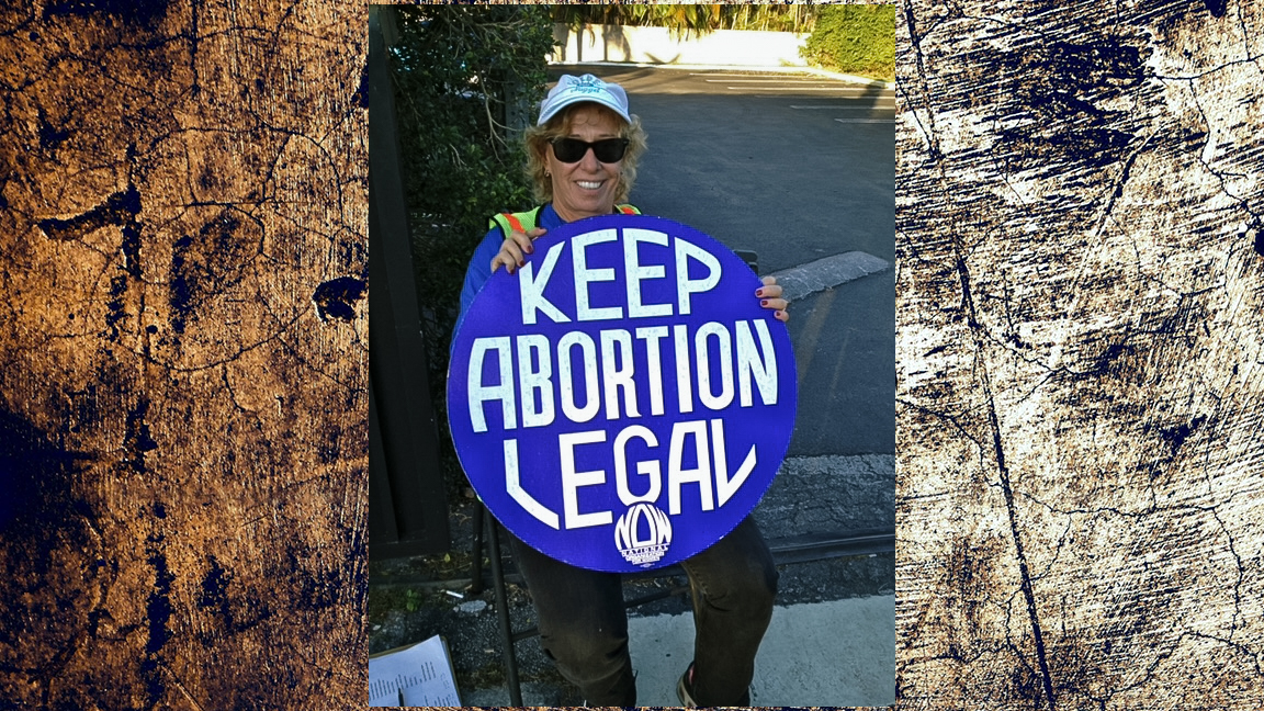 A person wearing sunglasses and a cap, holding a large blue circular sign that reads "KEEP ABORTION LEGAL" with the NOW (National Organization for Women) logo at the bottom. They are sitting outdoors on a sunny day.