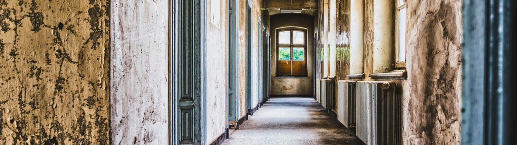 Long, abandoned hallway with peeling paint, cracked walls, and broken windows, leading to a closed wooden door with light streaming through at the far end.